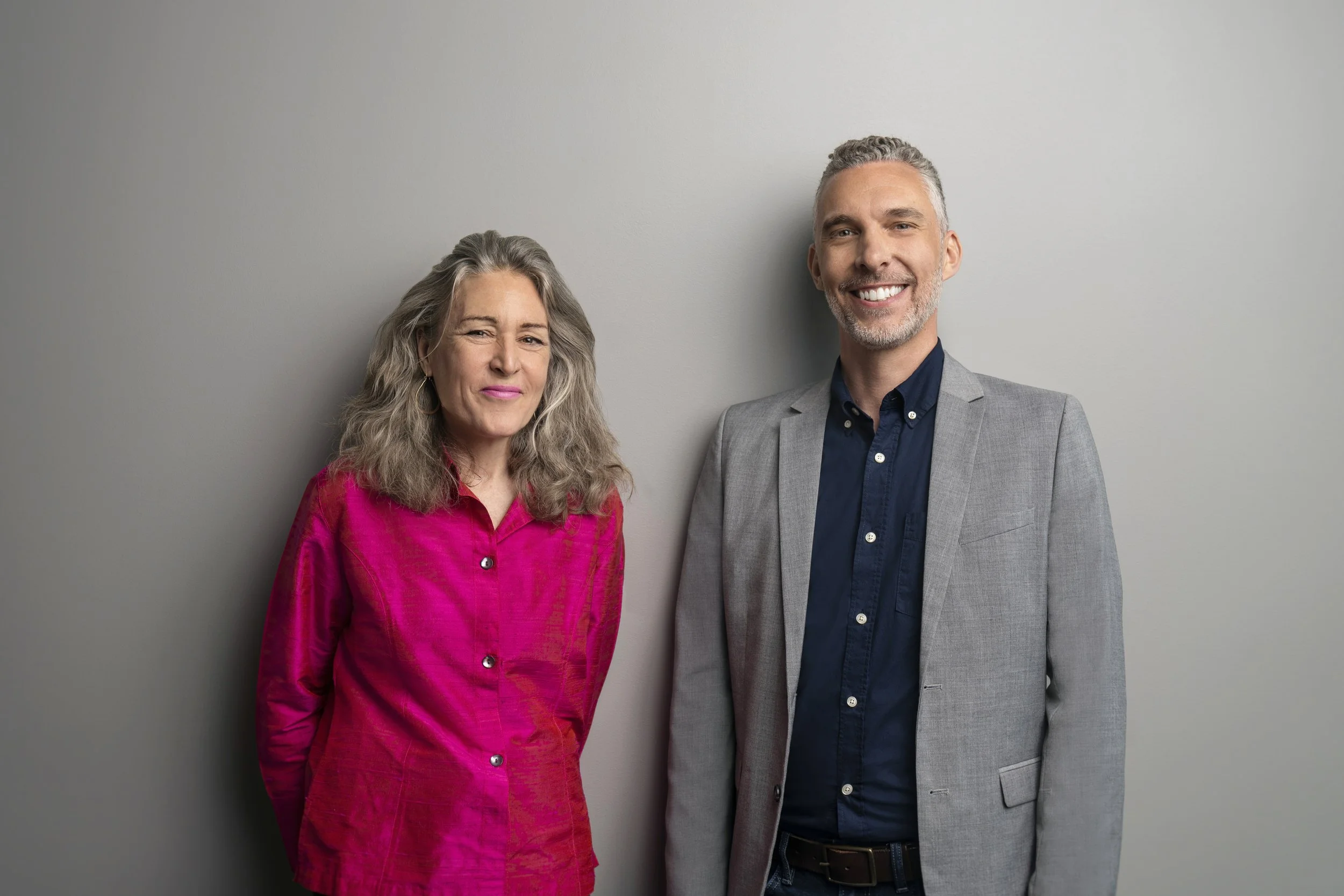 A woman and a man standing side by side against a plain gray wall, smiling at the camera.