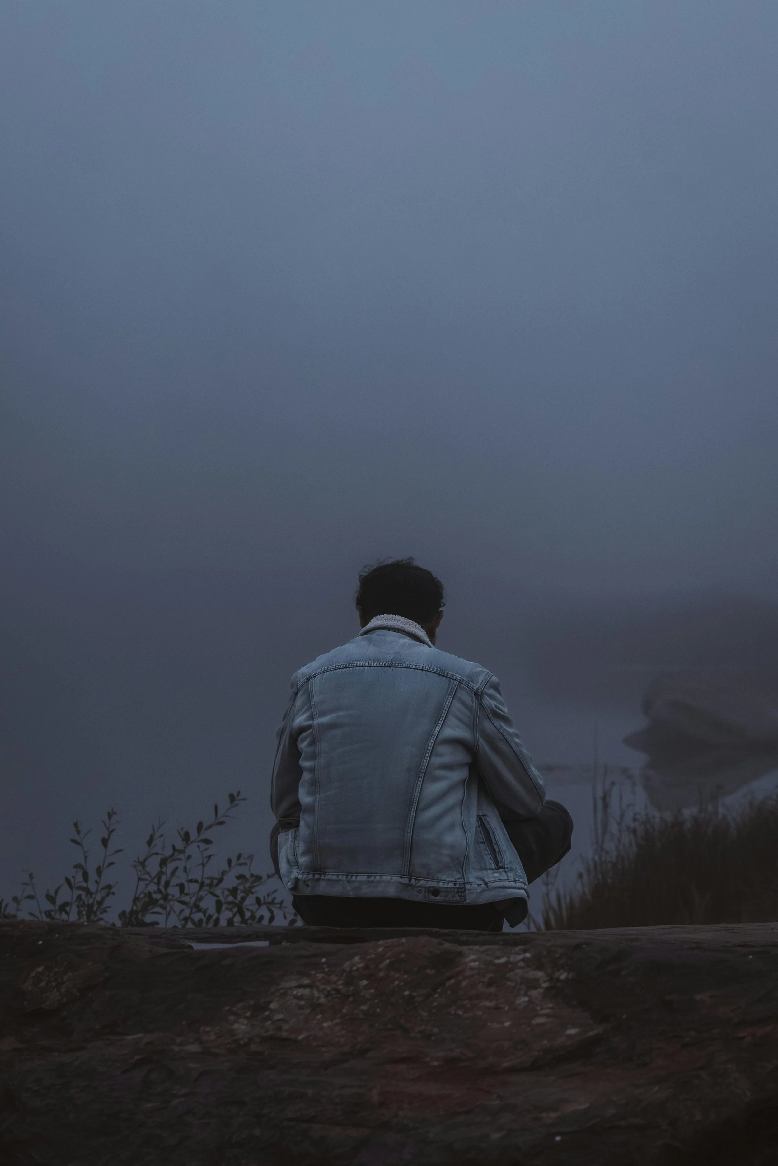 Man with dark hair wearing a light-colored denim jacket sitting outdoors during dusk, facing away from the camera, with a cloudy sky and horizon in the background.