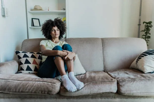 Young woman with curly hair sitting alone on a beige couch in a living room, looking thoughtful or sad, with decorative pillows and shelves in the background.