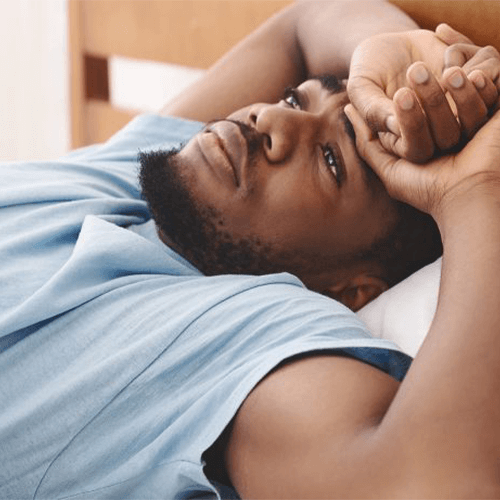 A man lying in bed with his eyes open, resting his head on his arm, wearing a light blue shirt.