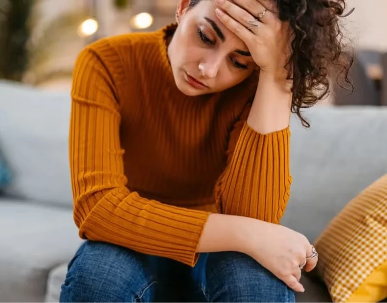 Woman sitting on a sofa with a worried or stressed expression, hand on forehead, in a cozy indoor setting.
