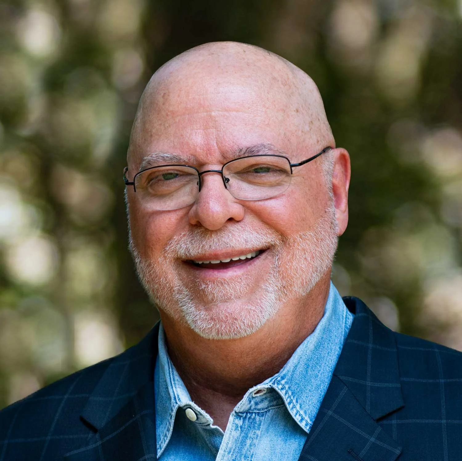 A smiling middle-aged man with glasses, a white beard, and a mustache wearing a dark blue checkered blazer over a light blue shirt, standing outdoors with a blurred green background.