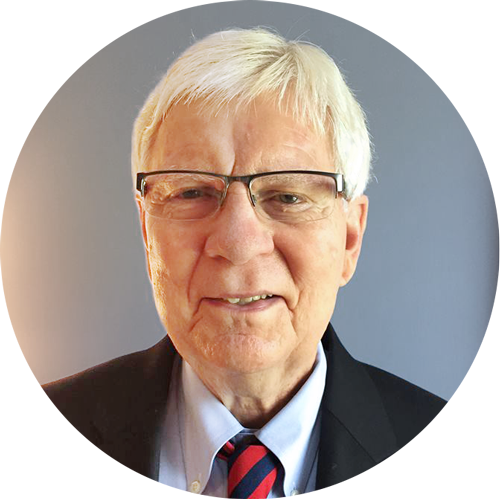 Portrait of an older man with gray hair, glasses, wearing a suit, white shirt, and a red and blue striped tie, standing in front of a blue background.
