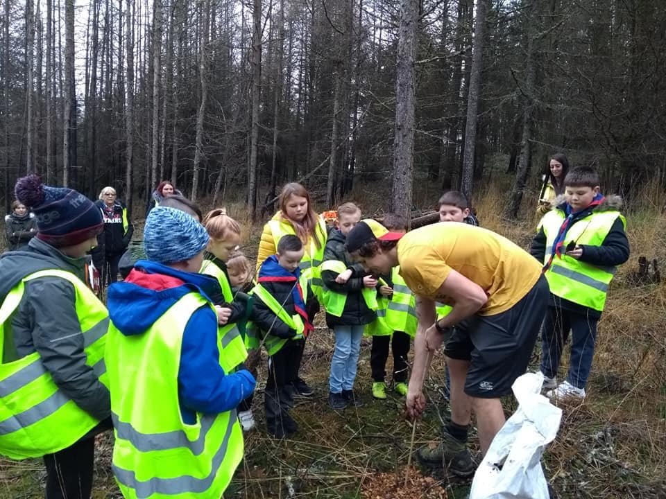 cub scouts and the leader in a forest setting all huddled around together as they are shown how to plant trees