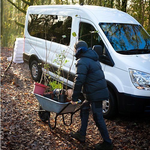 male wearing a jacket pushing a wheelbarrow with trees walking past a large white van