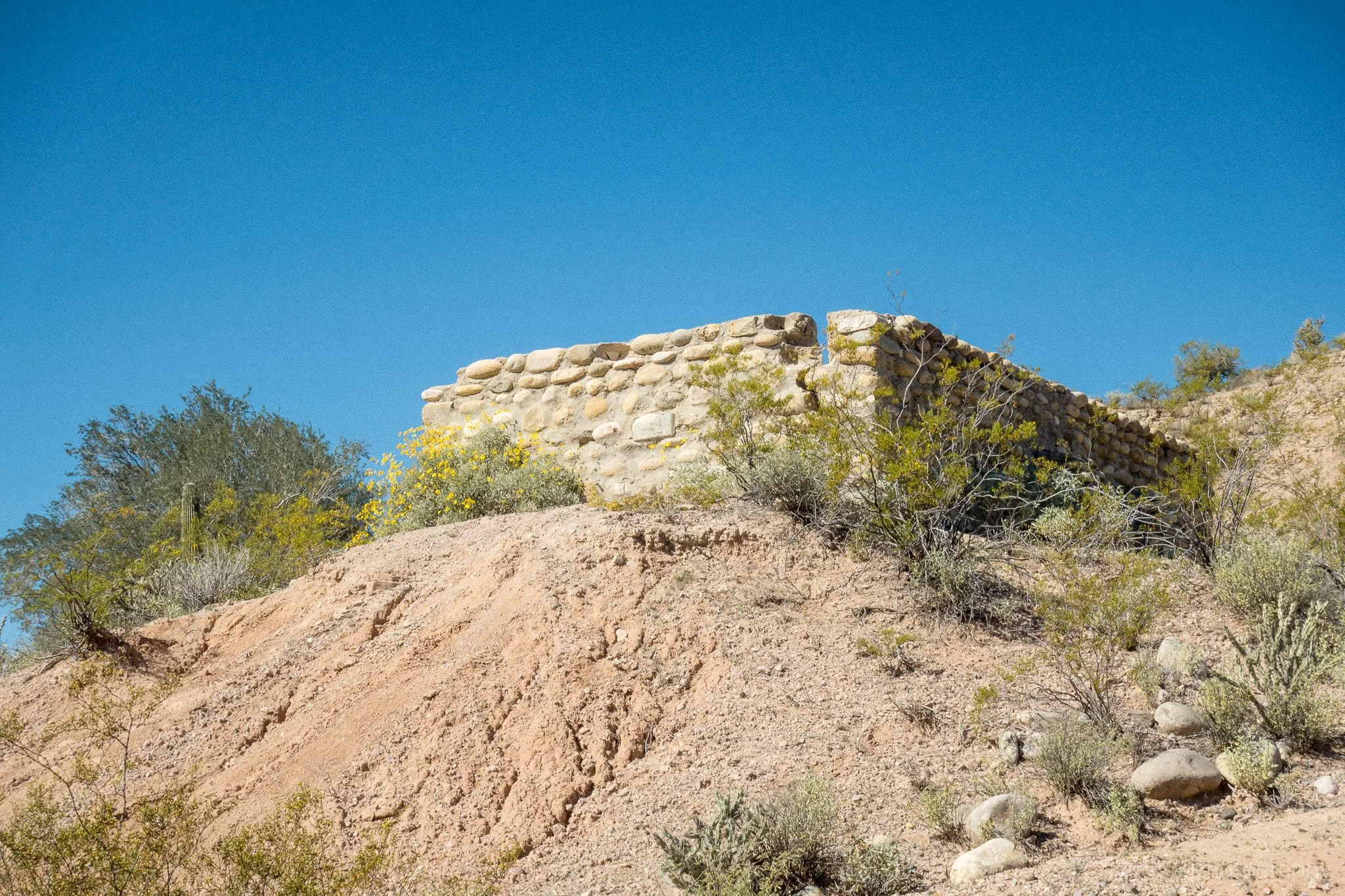 Stone wall structure in the Arizona desert surrounded by shrubs and sandy hillside terrain.