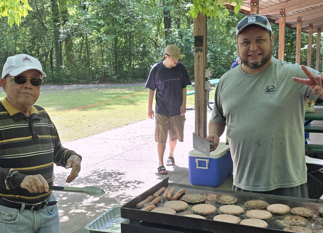 People grilling burger patties and hot dogs outdoors under a wooden shelter, with trees and greenery in the background.