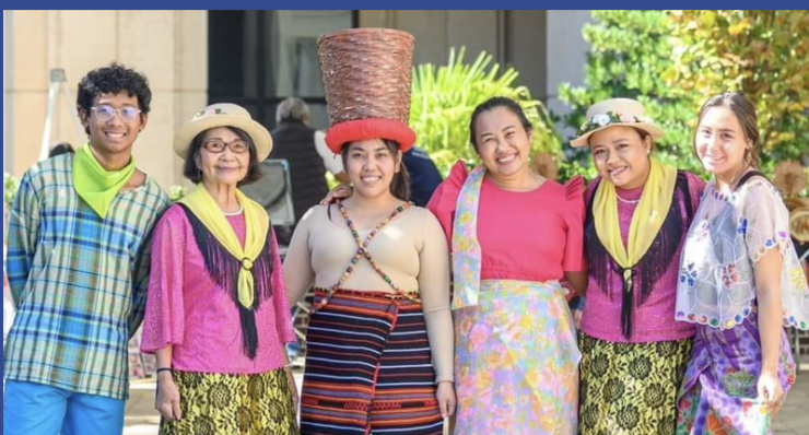 Group of six diverse women and one man smiling outdoors, dressed in colorful and traditional clothing, with one woman balancing a large basket on her head.