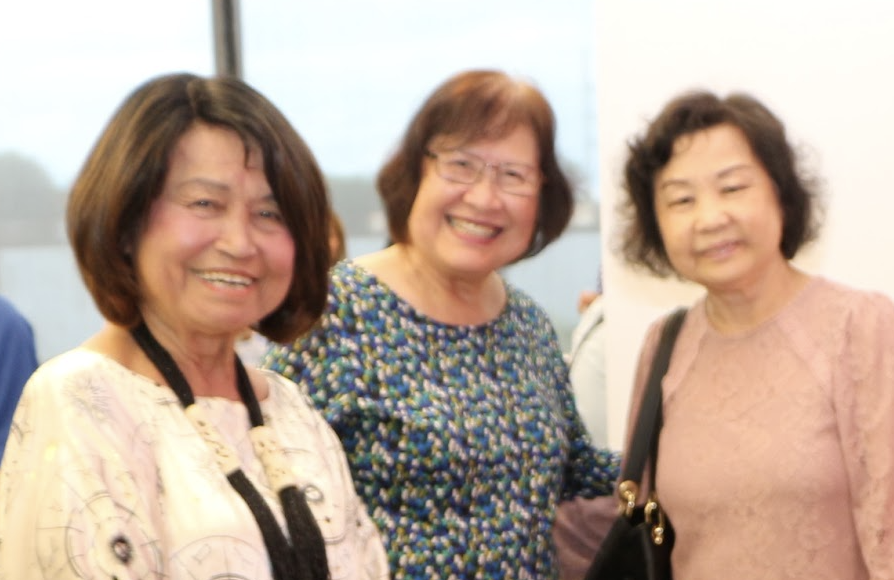 Three women smiling at a social gathering, standing indoors near a window.