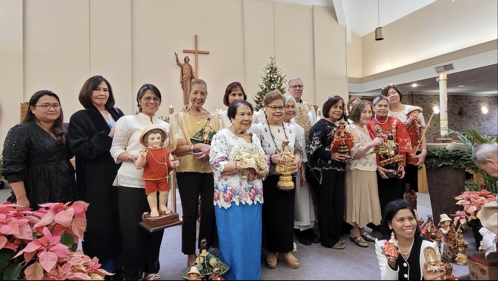 Group of people standing inside a church holding religious figurines and statues, with a Christmas tree and a crucifix on the wall behind them, decorated for Christmas.