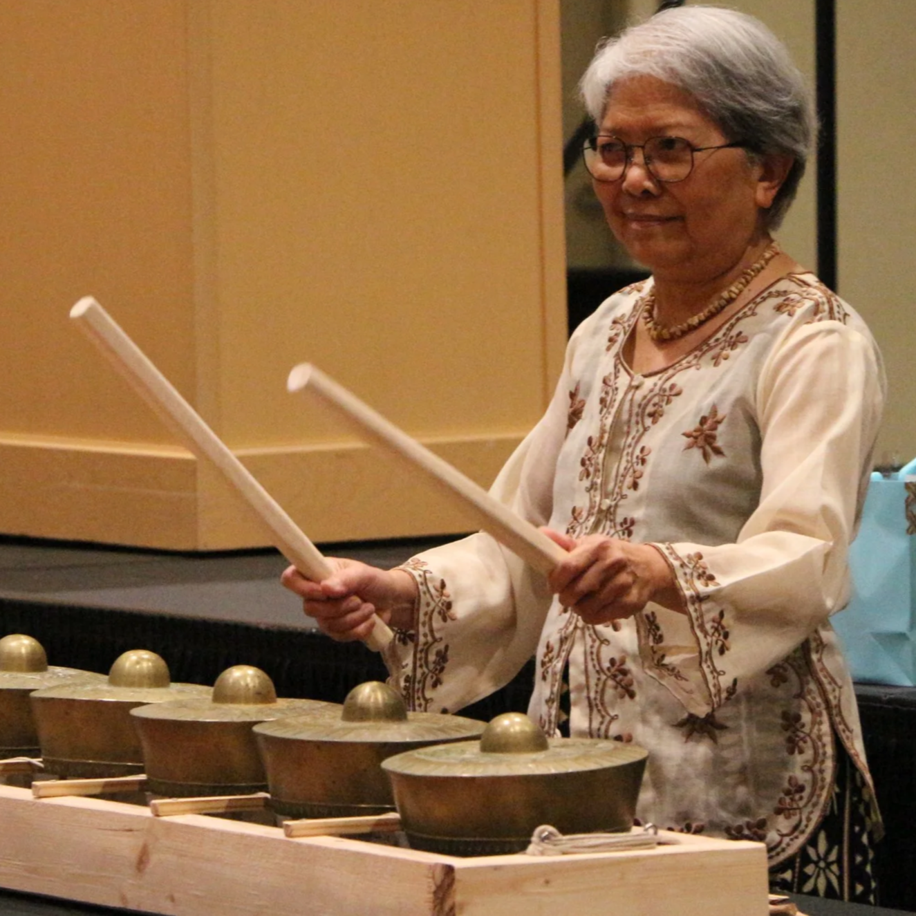 An elderly woman with white hair, glasses, and a traditional embroidered top playing a percussion instrument at an indoor event, with a percussion table in front and a wooden folding screen behind her.