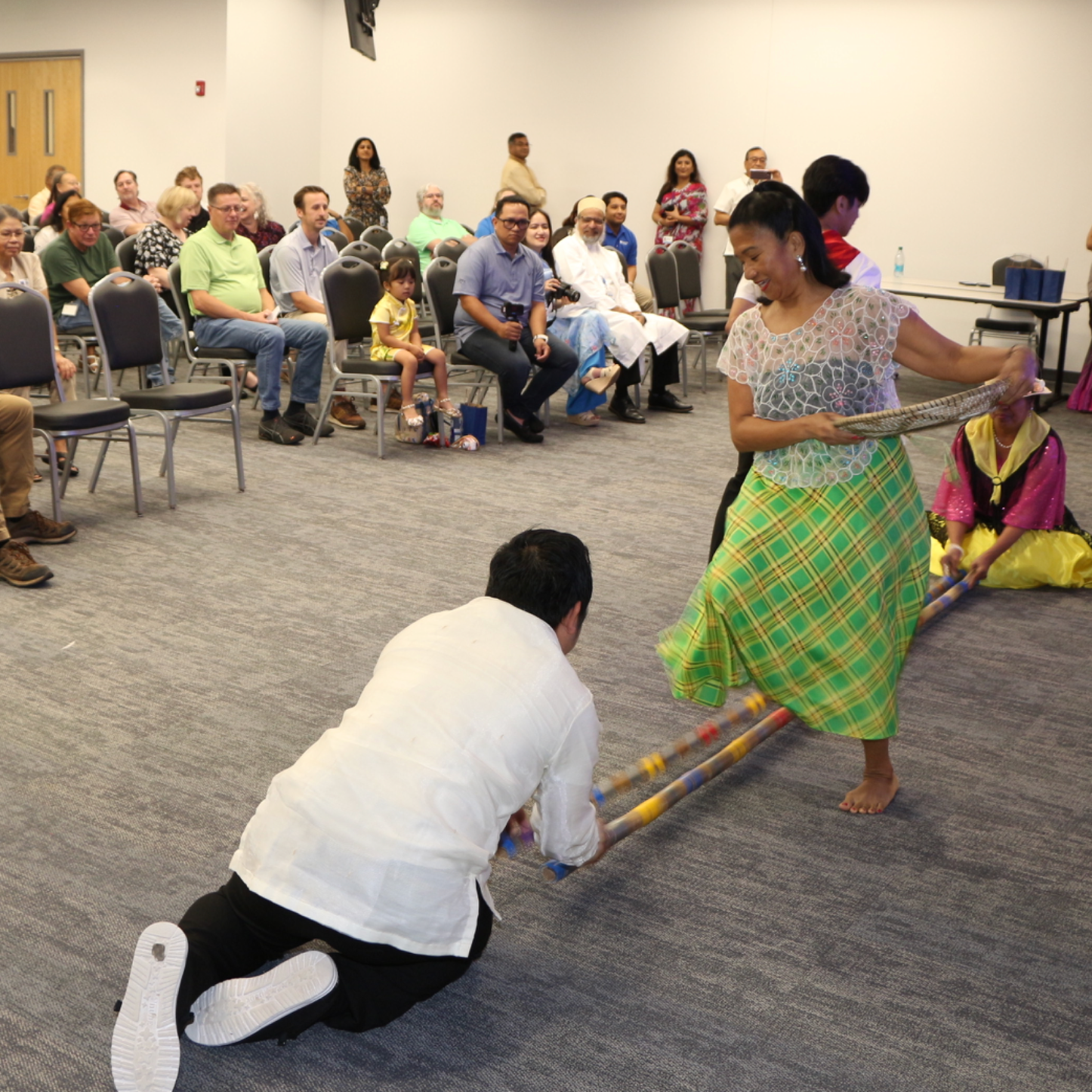 A diverse group of people seated in a conference room watching a performance. Two performers in traditional attire, one kneeling and the other standing, are engaging with a long stick as part of an activity.