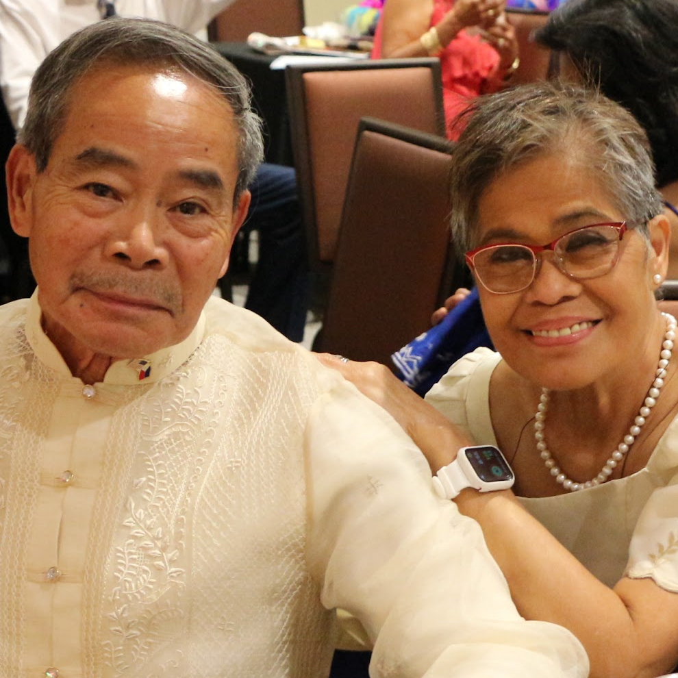 Older man and woman smiling at a banquet, dressed in traditional Filipino attire, with other guests in the background.