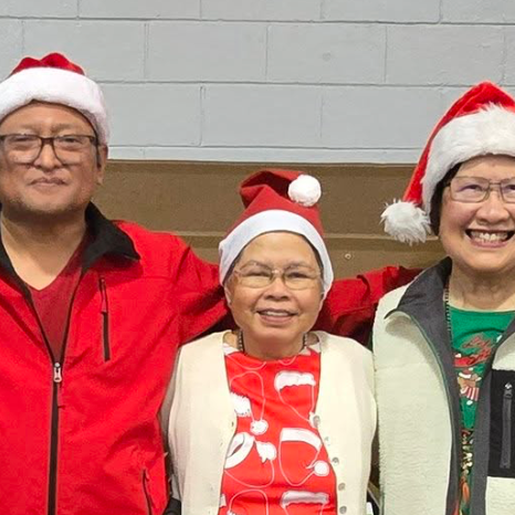 Three smiling people wearing Santa hats standing together indoors, two women and one man, dressed in colorful casual clothing.