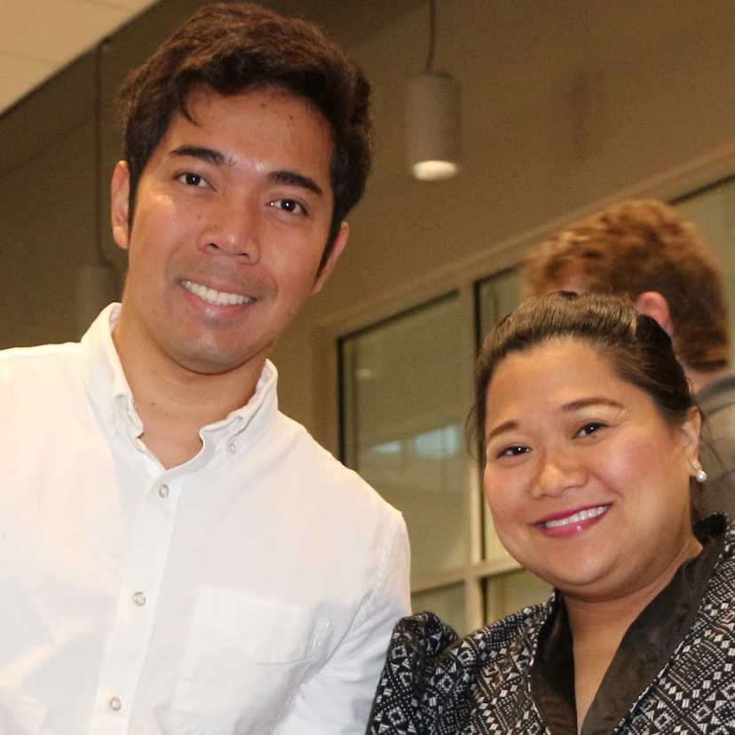 A young man with dark hair and a white button-up shirt smiling next to a woman with dark hair in a bun, wearing a black and white patterned blouse, both smiling at the camera in an indoor setting.