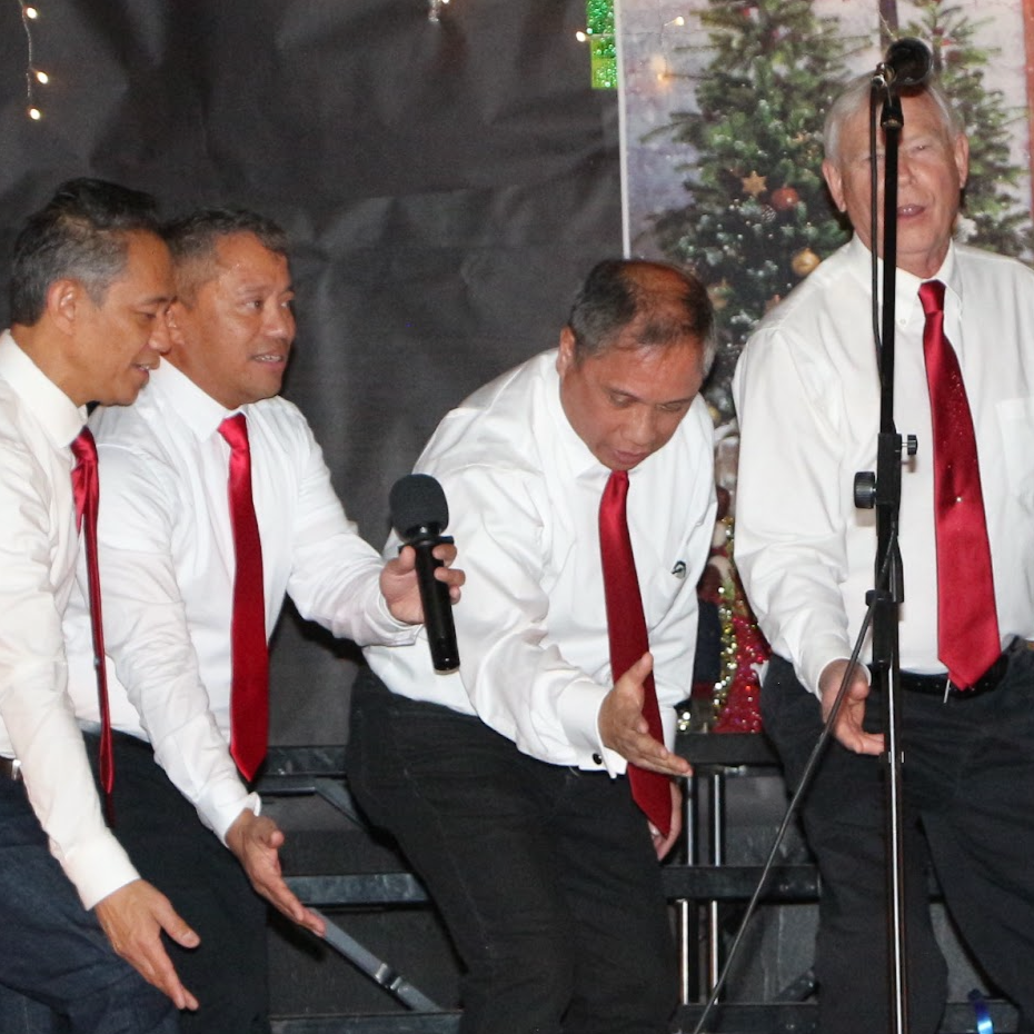 A group of people performing Christmas carol singing on stage with Christmas decorations in the background.