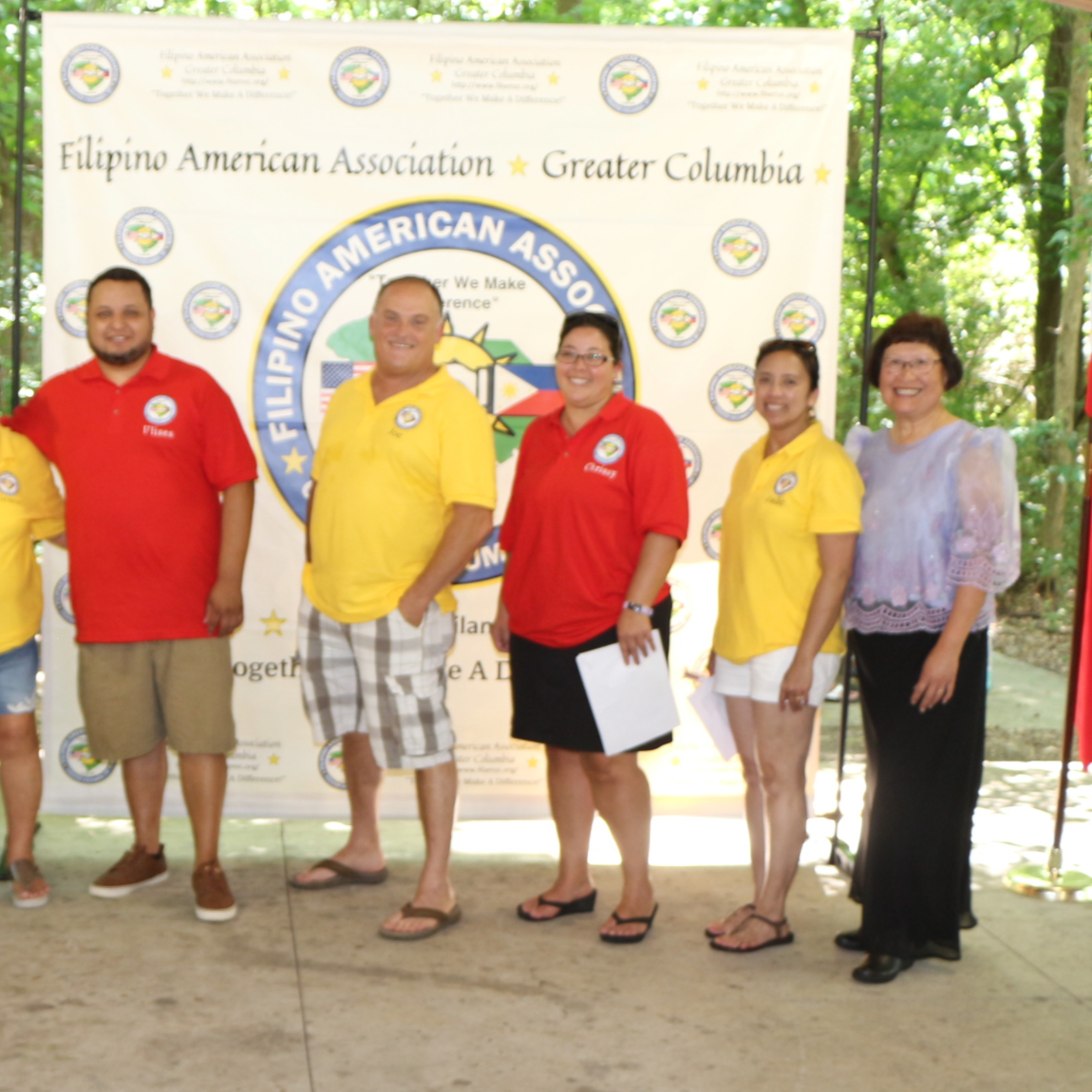 A group of ten people standing outdoors in front of a large banner for the Filipino American Association of Greater Columbia, with American and Filipino flags on either side. The group is diverse in age and gender, with some wearing yellow or red shi