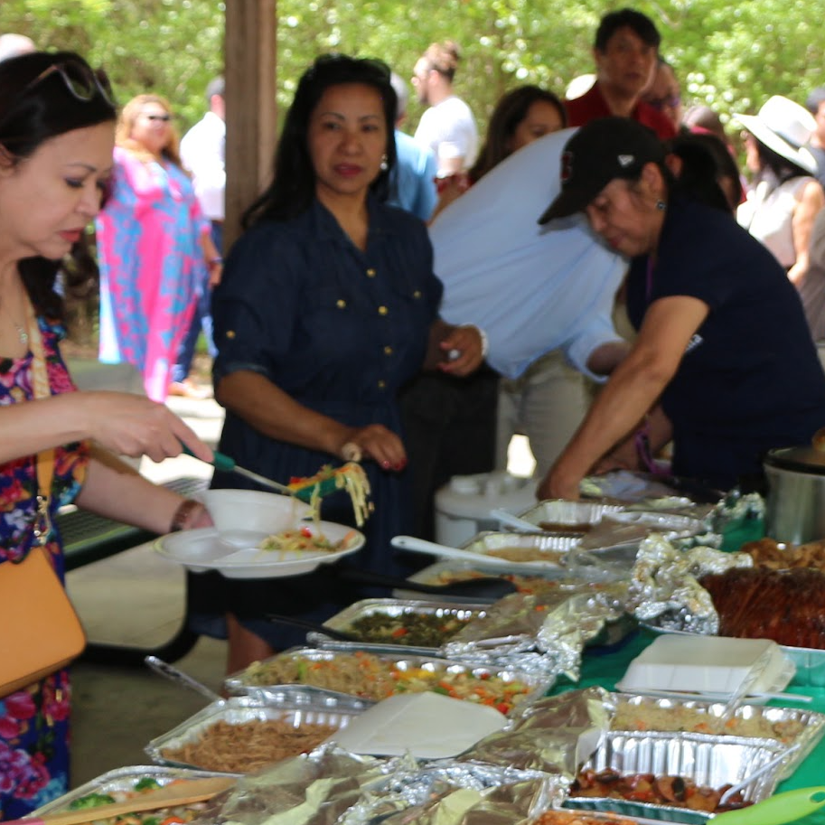 People serving themselves food at an outdoor buffet with trays of various dishes.
