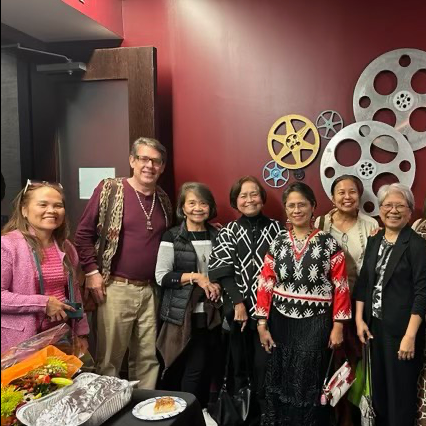 Group of eleven diverse adults, mostly women, standing together at a social gathering, with some holding drinks and smiling, against a red wall decorated with large film reel wall art, a table with snacks and food items visible in the foreground.