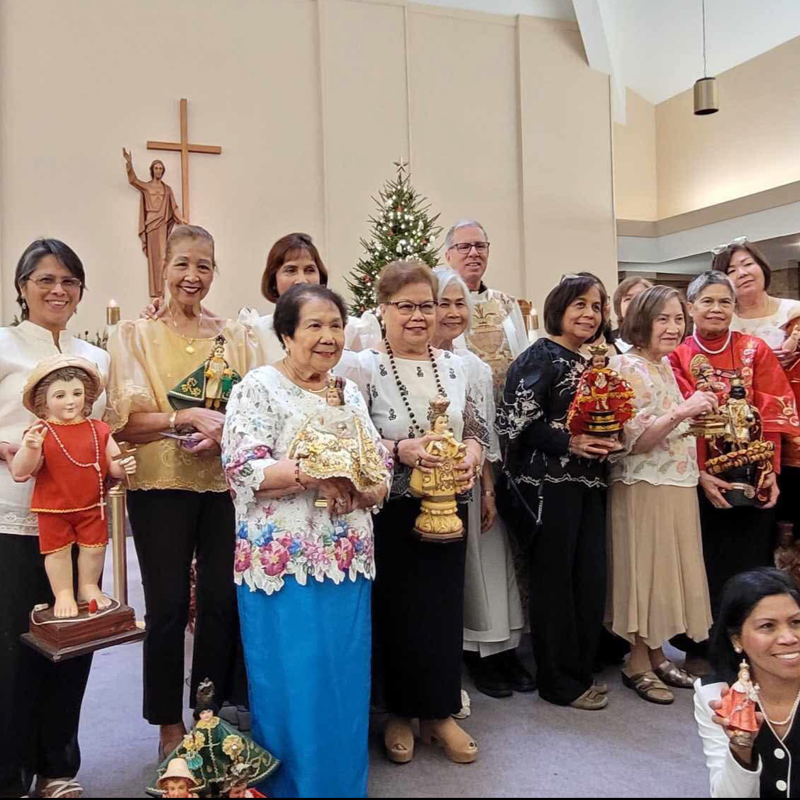 Group of people standing inside a church holding religious figurines and statues, with a Christmas tree and a crucifix on the wall behind them, decorated for Christmas.