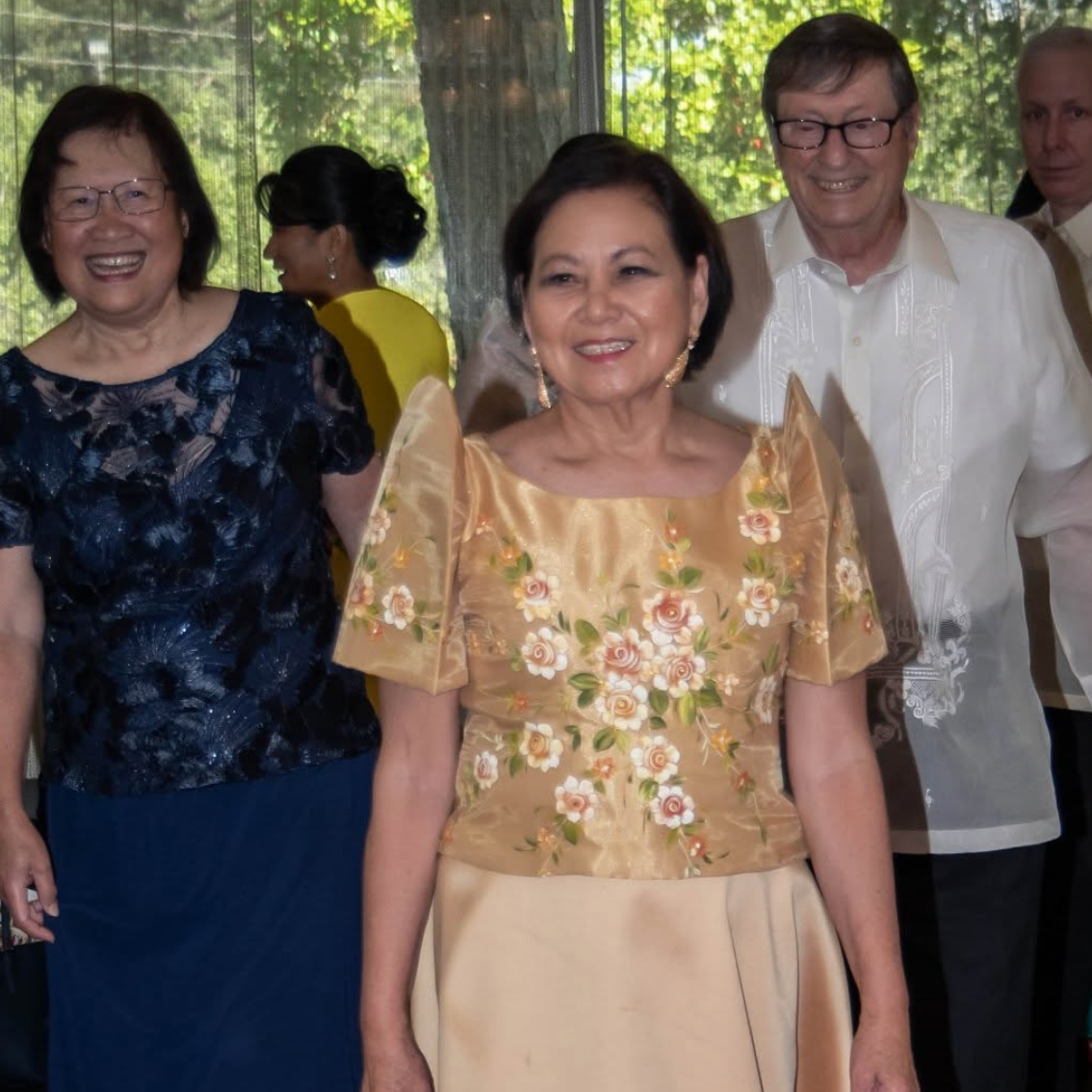 A group of well-dressed people at a formal event, with two women in traditional Filipino attire and two men in formal suits, standing on a red carpet with a green outdoor background visible through a window.