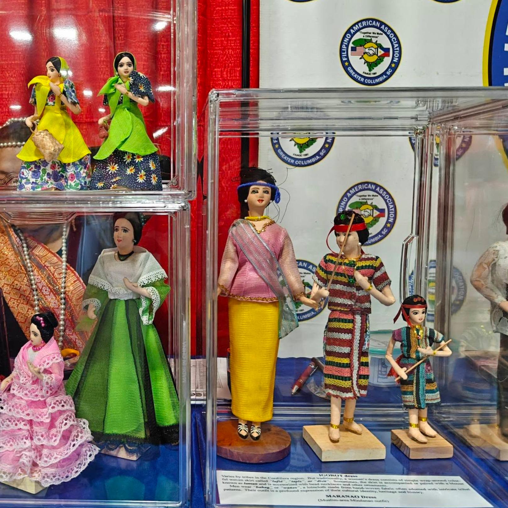 Display of traditional Filipino dolls dressed in colorful clothing on a clear plastic shelf at a cultural exhibit.
