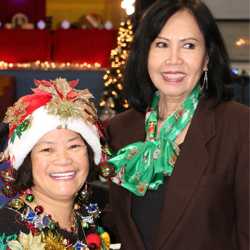 Two women celebrating Christmas, one wearing a Santa hat decorated with ornaments and the other in a brown blazer with a green festive scarf, surrounded by Christmas decorations and a decorated tree in the background.
