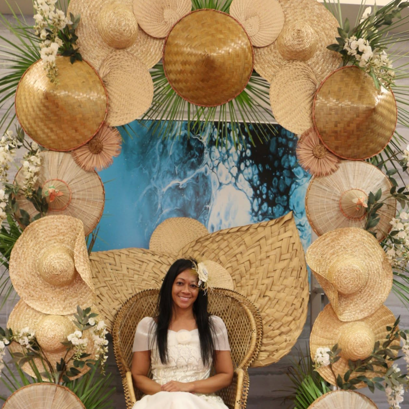 A woman in a white dress sits on a wicker chair in front of a decorative backdrop of straw hats and green foliage, with a blue abstract painting behind her.