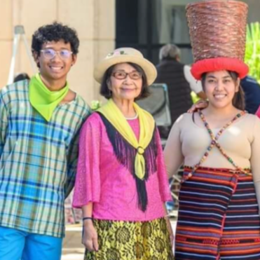 Group of six diverse women and one man smiling outdoors, dressed in colorful and traditional clothing, with one woman balancing a large basket on her head.