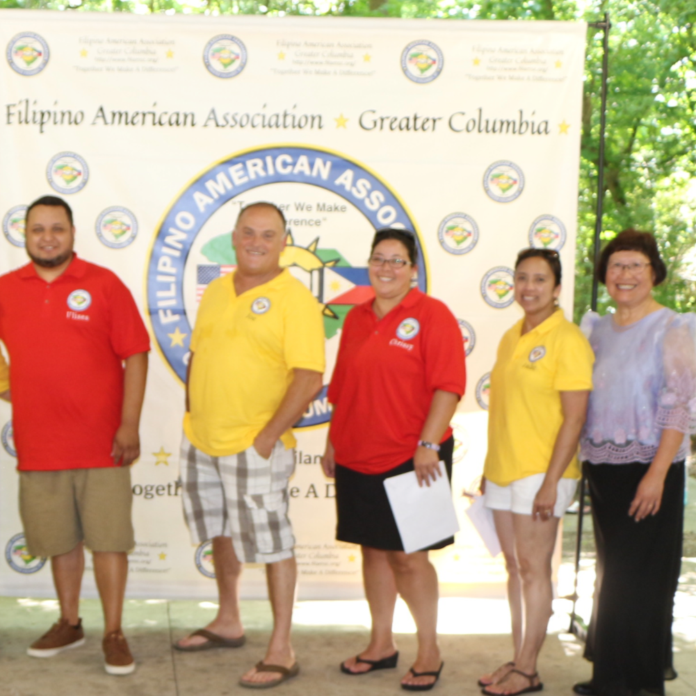 A group of ten people standing outdoors in front of a large banner for the Filipino American Association of Greater Columbia, with American and Filipino flags on either side. The group is diverse in age and gender, with some wearing yellow or red shi