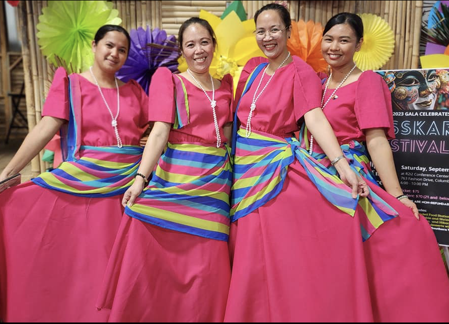 Four women in pink traditional dresses with colorful striped sashes, standing side by side, smiling at an event with colorful paper decorations in the background.