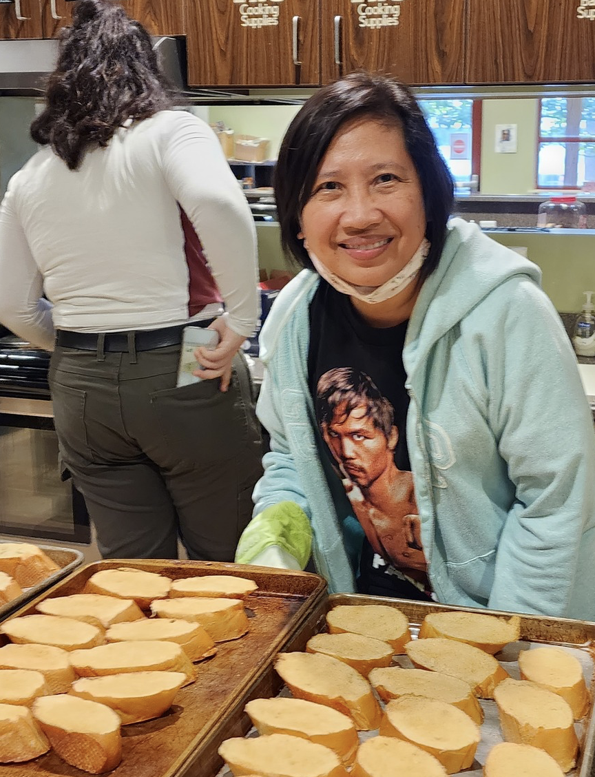 Smiling woman in a light hoodie and a Bob Marley t-shirt, wearing a face mask, preparing sliced bread on a baking sheet in a kitchen.
