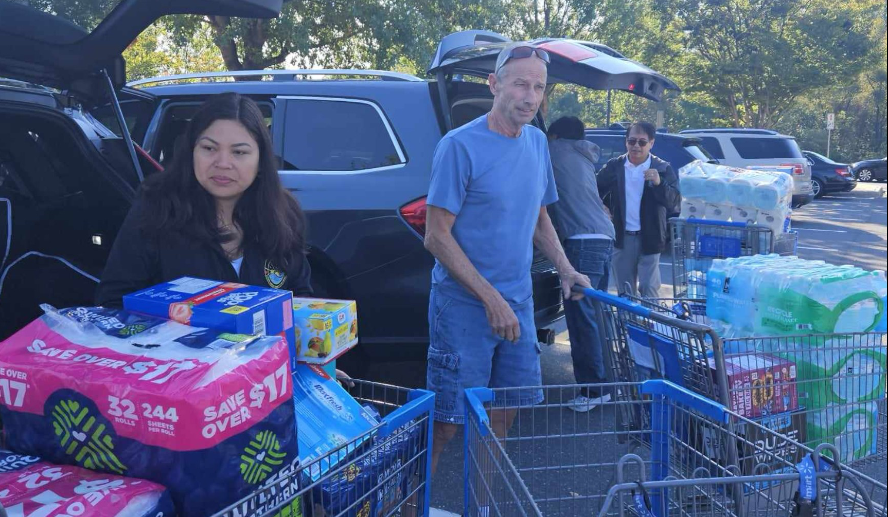People at a parking lot participating in a food or supply distribution event, with carts filled with packages of supplies and bottled water, cars in the background, and trees surrounding the area.