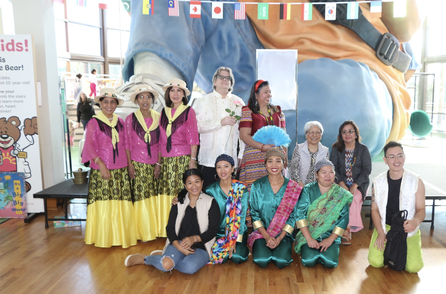 Group of people in cultural attire posing in front of a large cartoon bear sculpture at an indoor event.