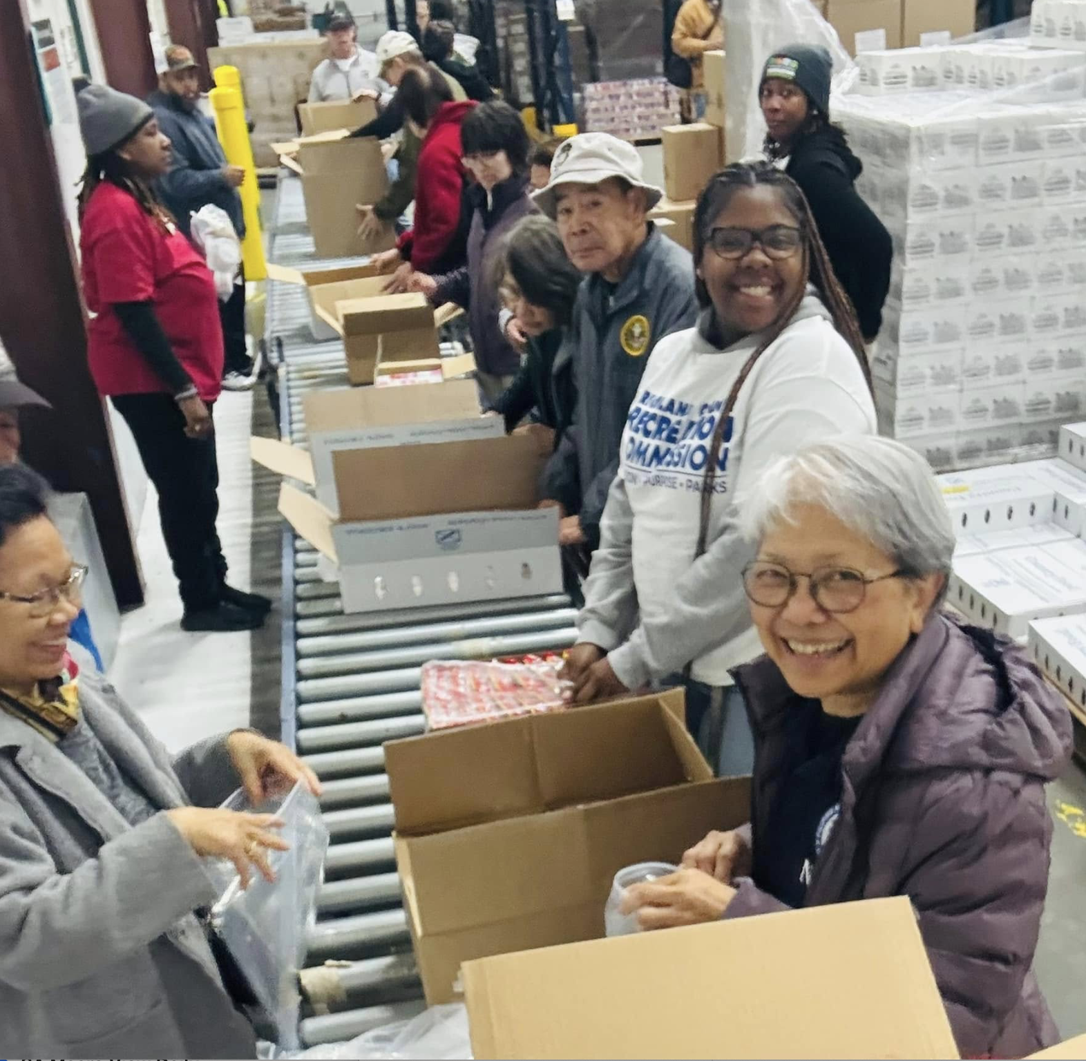 People volunteering at a warehouse, sorting and packing items for distribution, with several individuals smiling and engaging in the activity.