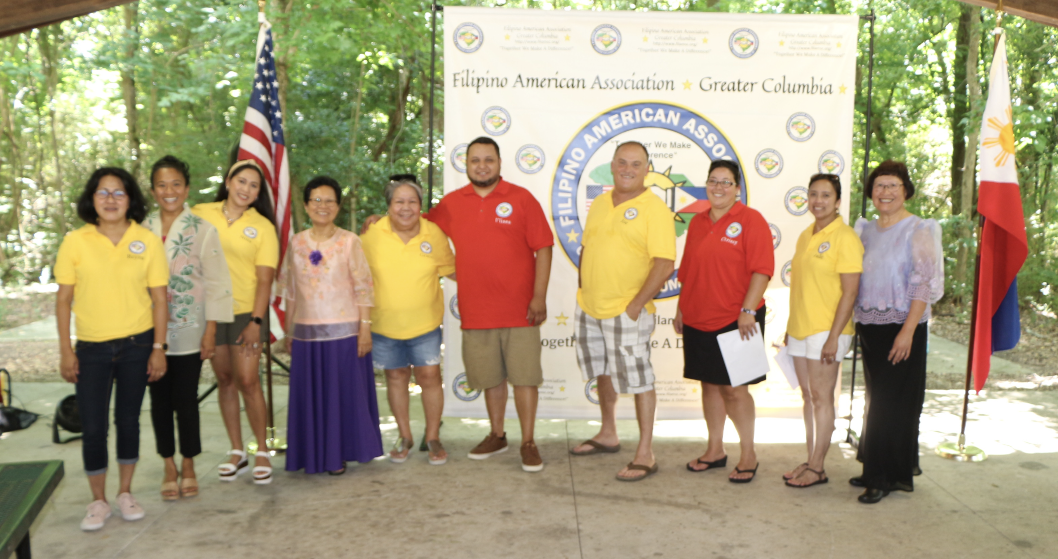 A group of ten people standing outdoors in front of a large banner for the Filipino American Association of Greater Columbia, with American and Filipino flags on either side. The group is diverse in age and gender, with some wearing yellow or red shi