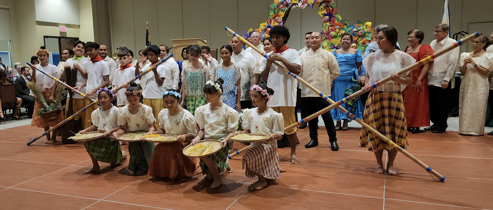 Group of people in traditional Filipino attire performing a cultural dance on stage, with some holding bamboo poles and others holding baskets or fans, and a colorful floral arch in the background.