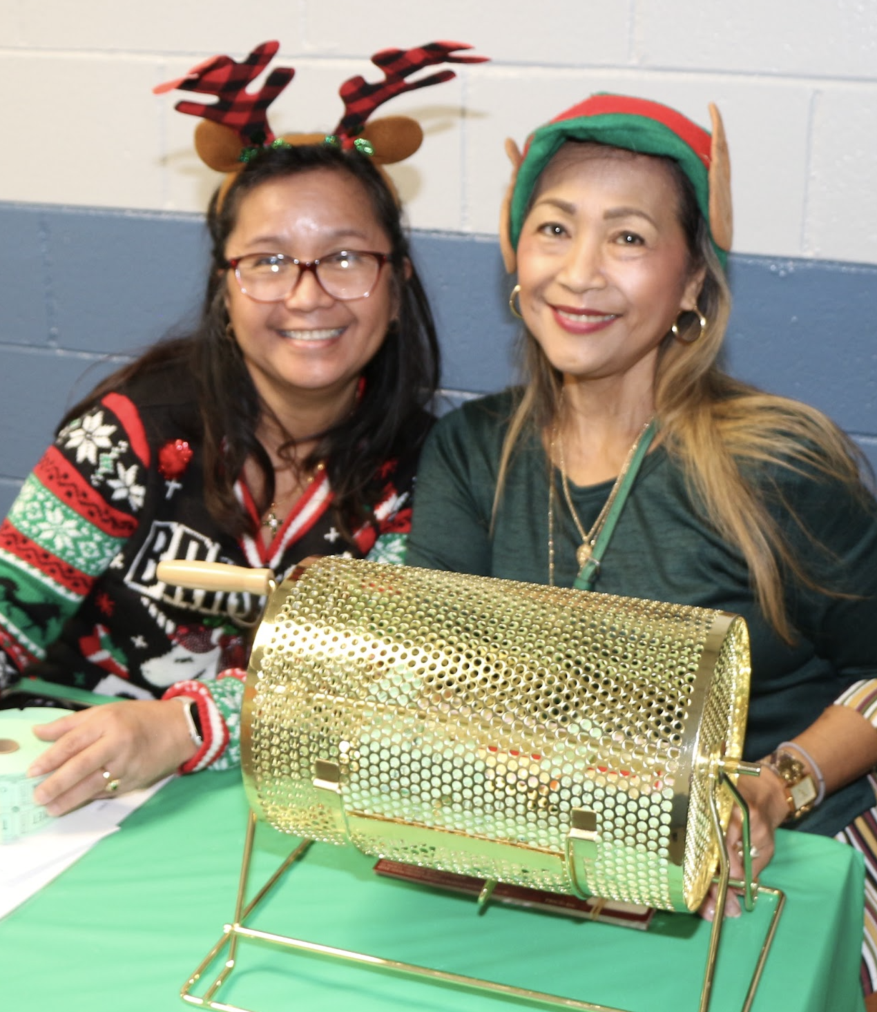 Two women in Christmas sweaters and festive accessories smiling at a holiday event, with a gold decorative raffle drum and green tablecloth in front of them.