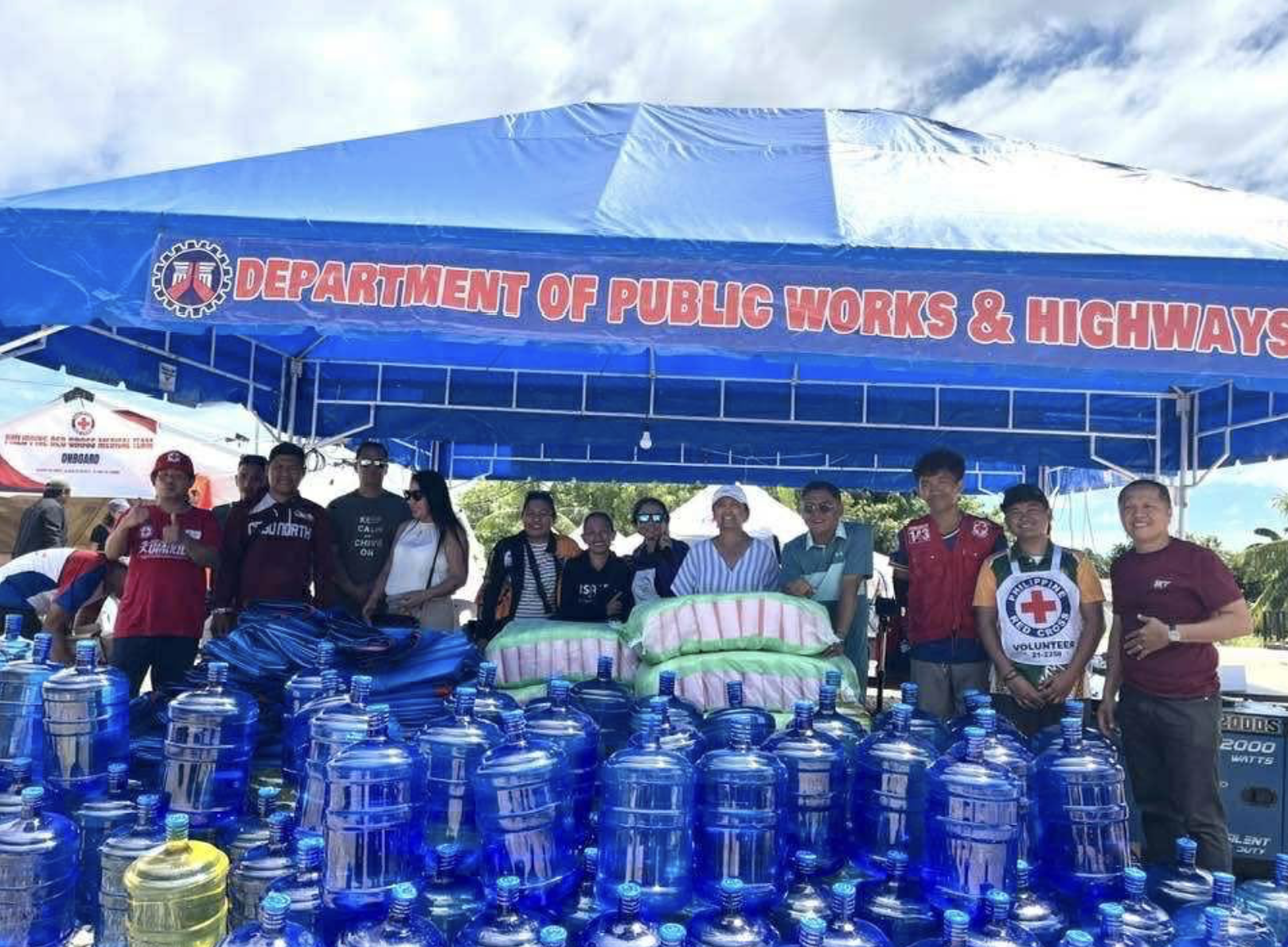 A group of people standing under a blue canopy with a sign that reads 'Department of Public Works & Highways.' In front of them are numerous bottles of water and some packs of supplies, with some individuals wearing volunteer vests and shirts.