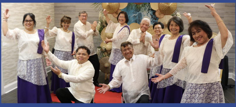 Group of nine people, mostly women and two men, dressed in traditional Filipino attire, posing together at a celebration or event with a decorated backdrop.