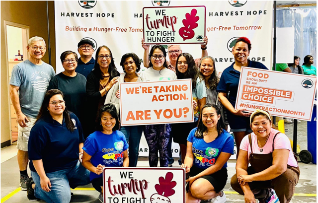 Group of people standing and kneeling at a hunger action event, holding signs that promote fighting hunger and taking action, with a banner reading 'Harvest Hope' in the background.