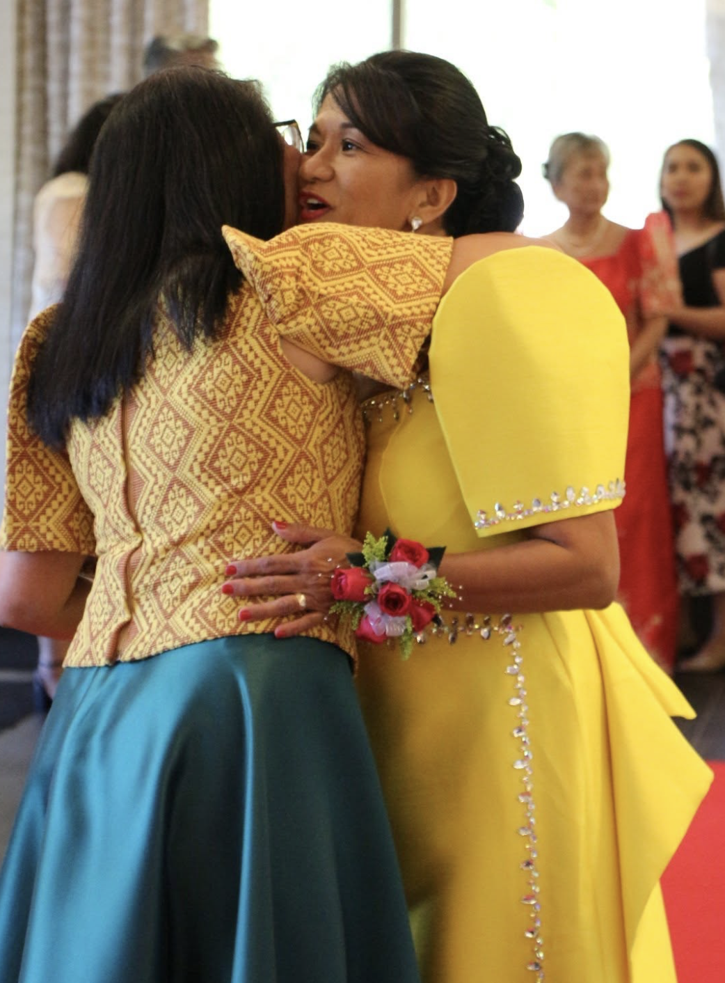 Two women hugging at a celebration, one wearing a yellow dress with floral embellishments and a corsage, the other wearing a patterned top and teal skirt, surrounded by other women in colorful dresses.