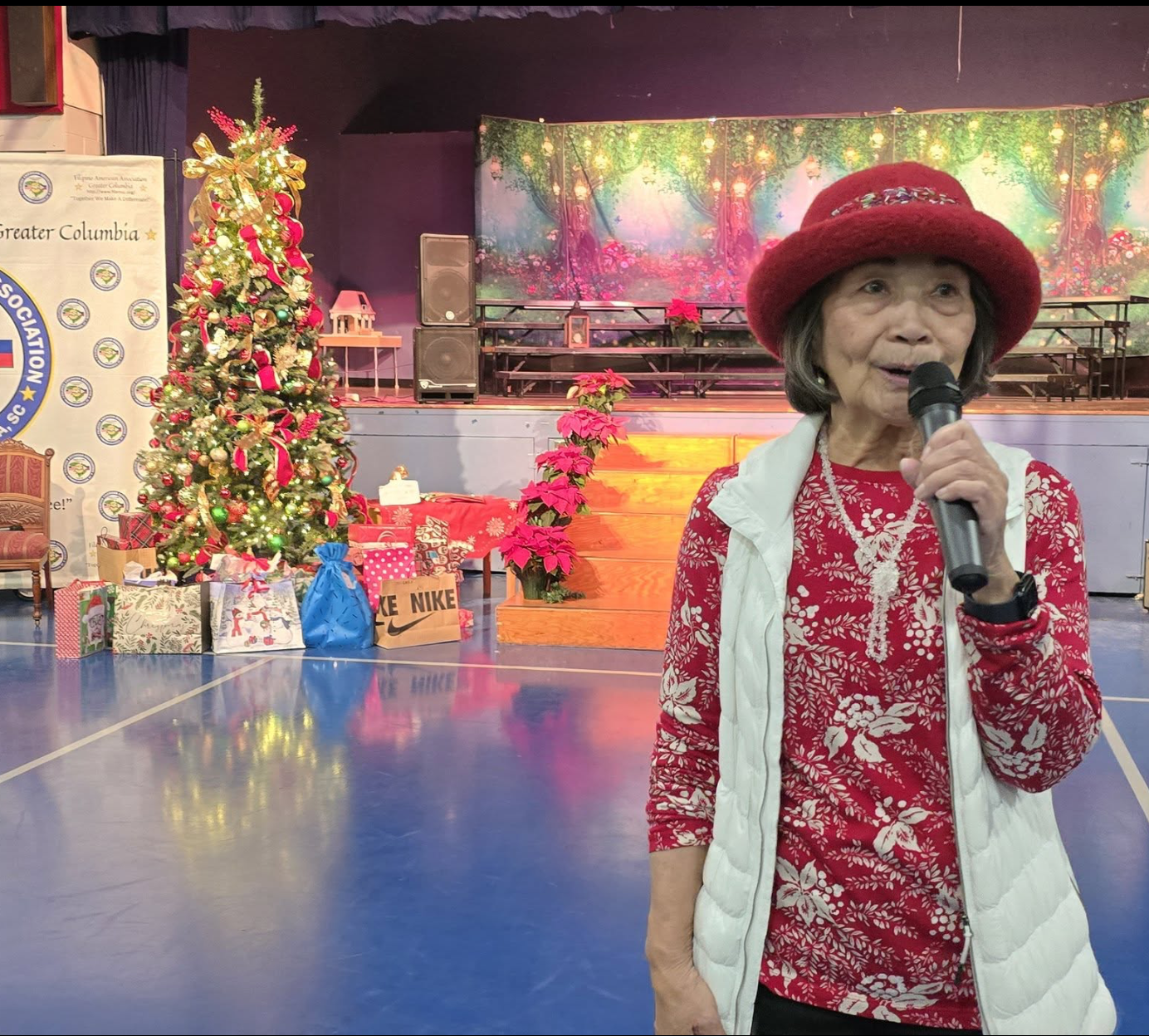 A woman in a red hat and red floral shirt speaking into a microphone at a Christmas event, with a decorated Christmas tree, wrapped gifts, and poinsettia plants in the background.