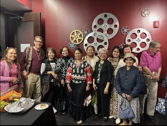 Group of eleven diverse adults, mostly women, standing together at a social gathering, with some holding drinks and smiling, against a red wall decorated with large film reel wall art, a table with snacks and food items visible in the foreground.