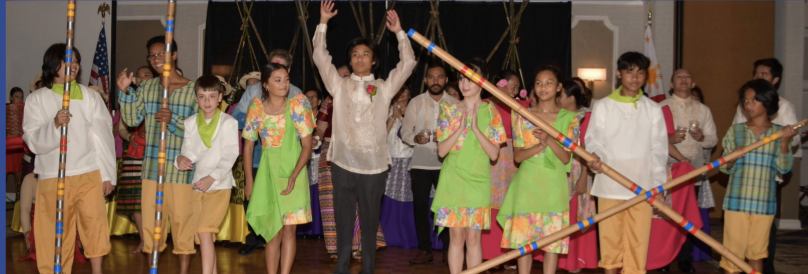 Group of people, including children and adults, participating in a cultural celebration or performance with decorated sticks, some in traditional attire, inside a room with American flags and a stage.