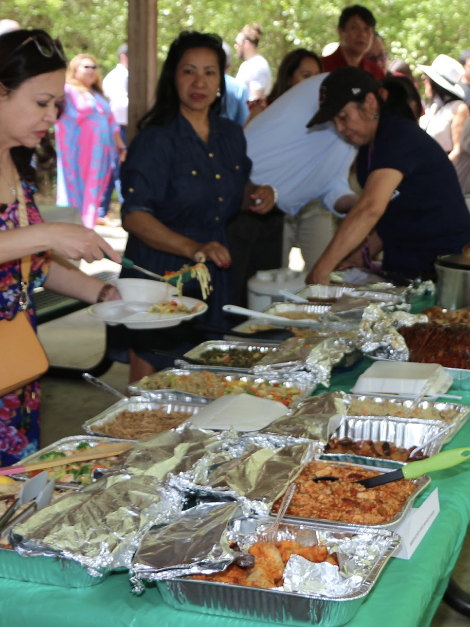 People serving themselves food at an outdoor buffet with trays of various dishes.