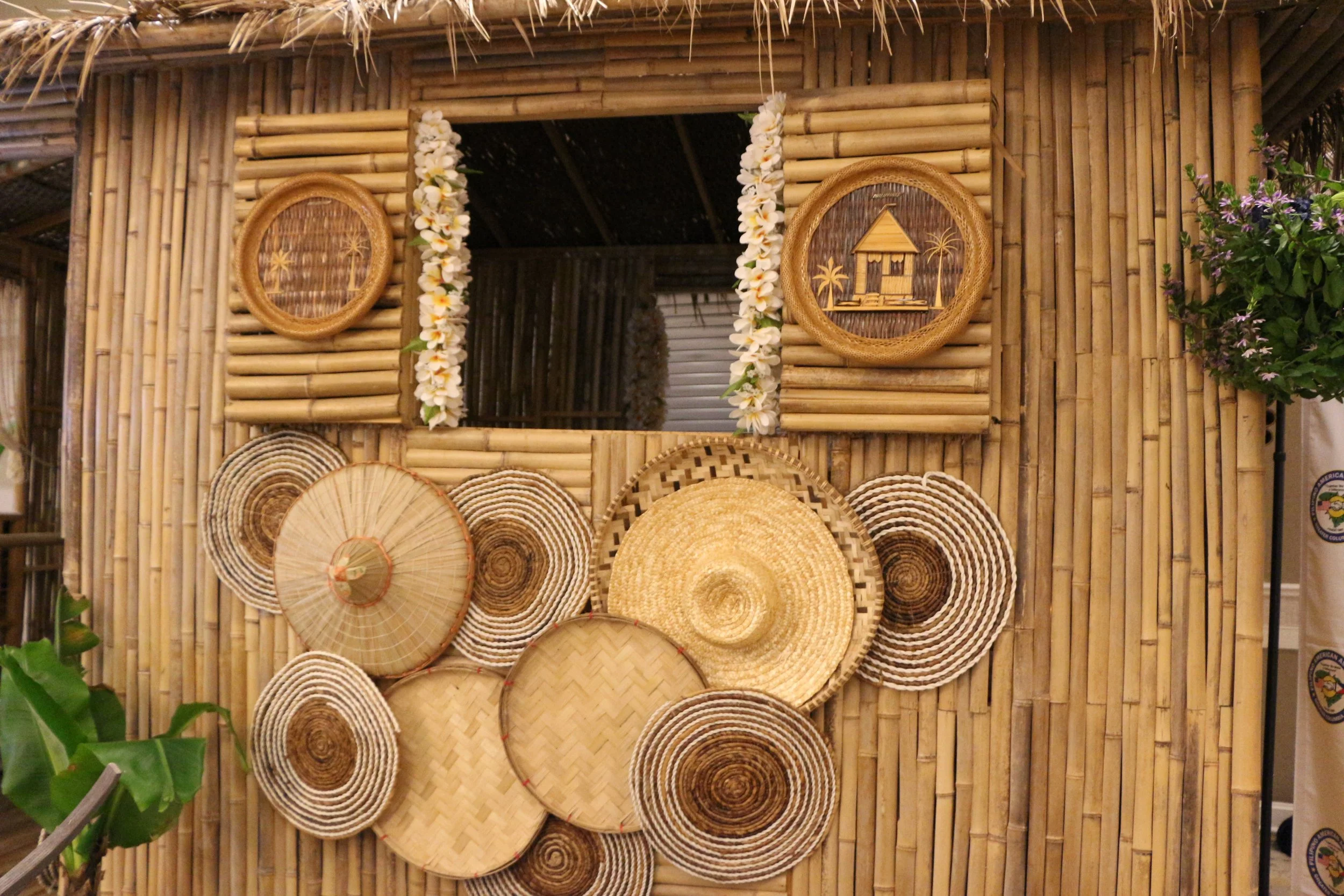 Display of woven baskets, hats, and decorative items on a bamboo wall with floral garland and potted plants nearby.