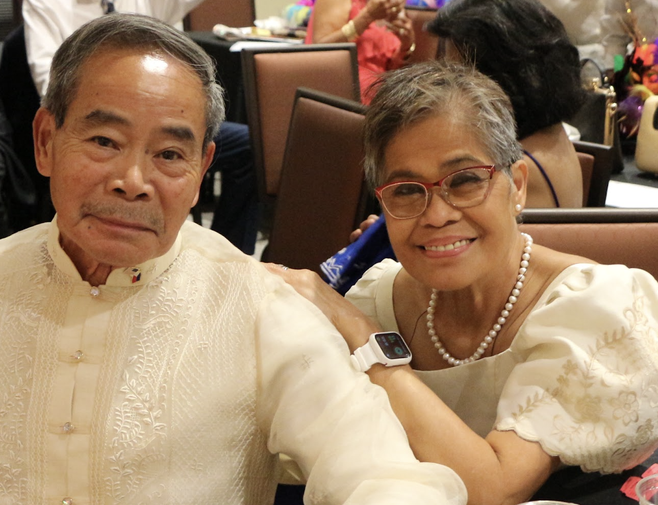 An elderly man and woman smiling at a banquet, both wearing traditional cream-colored Filipino attire, with the woman wearing glasses and a pearl necklace.