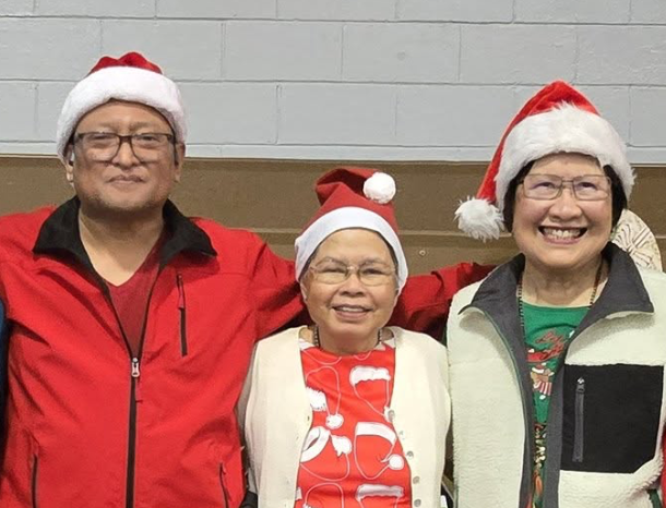Three smiling people wearing Santa hats standing together indoors, two women and one man, dressed in colorful casual clothing.