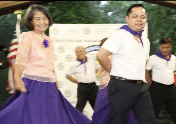 Elderly woman and young man dancing together outdoors, with others in the background, at a cultural event or gathering.